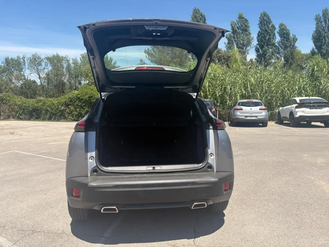 Vue arrière d’un Peugeot 2008 Hybrid 145 gris 2025 avec coffre ouvert et feux arrière allumés, en extérieur.