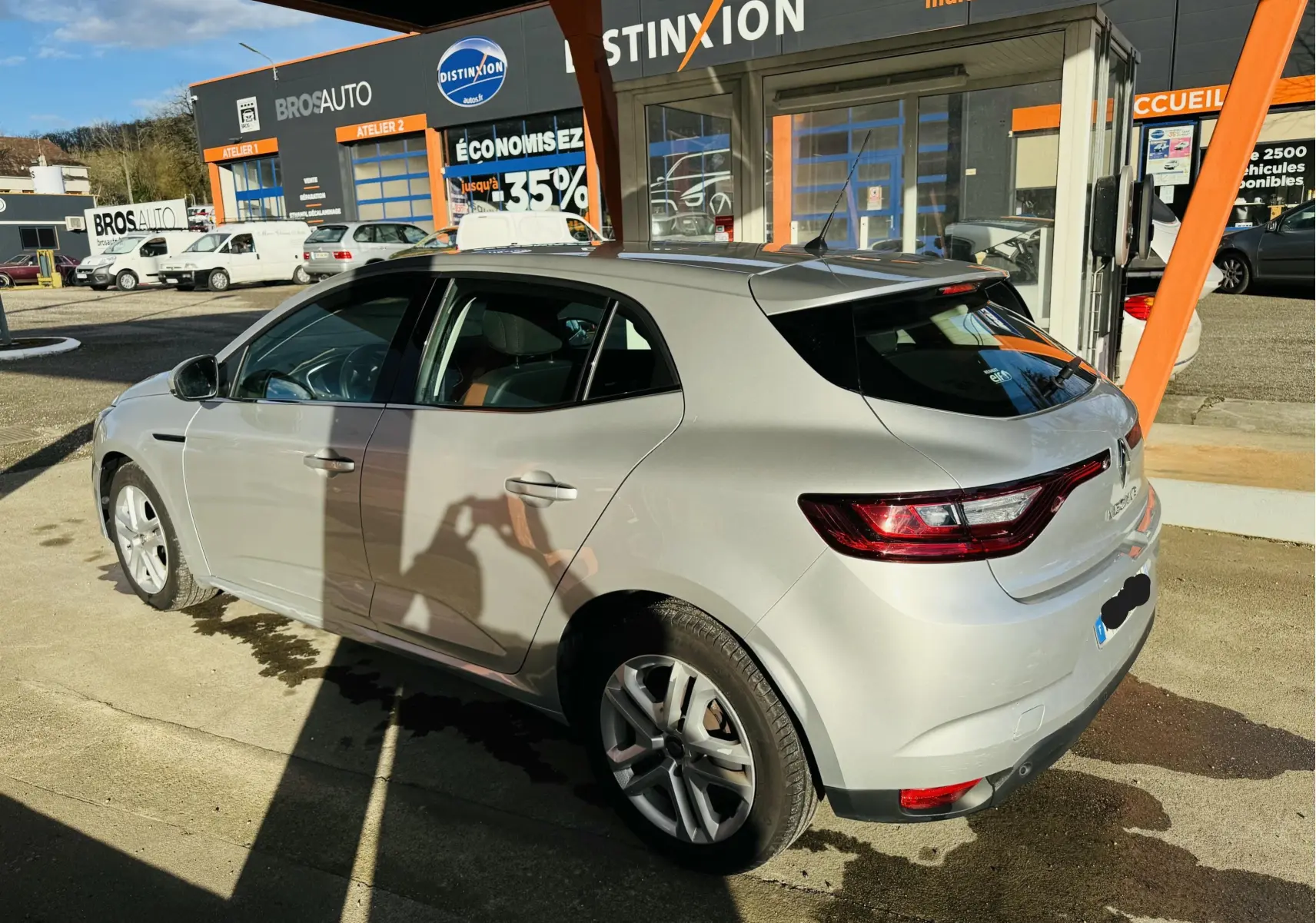 Vue 3/4 arrière droite d'une Renault Mégane IV berline gris métallisé stationnée devant un garage multimarque.