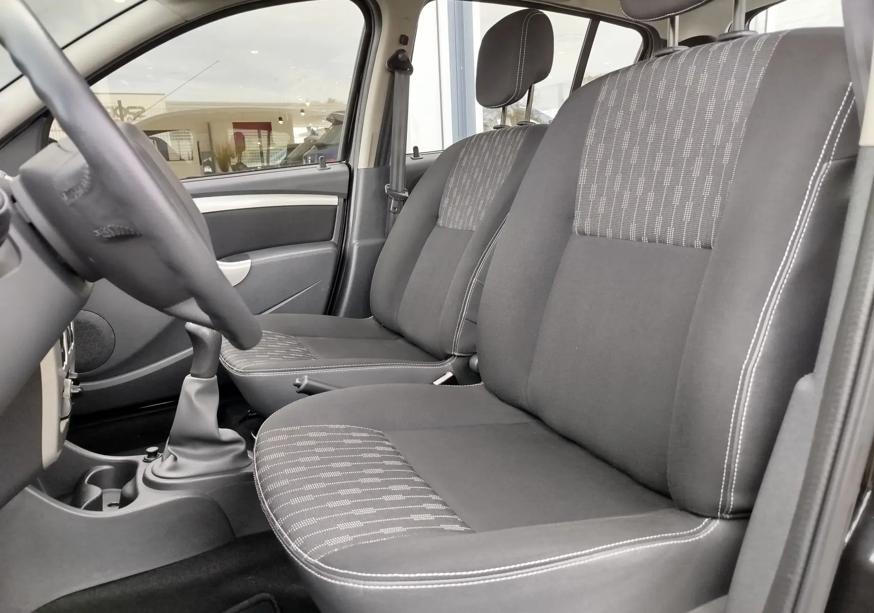 Intérieur avant de la Dacia Sandero noire 2011, vue côté gauche montrant sièges tissu gris avec surpiqûres blanches.