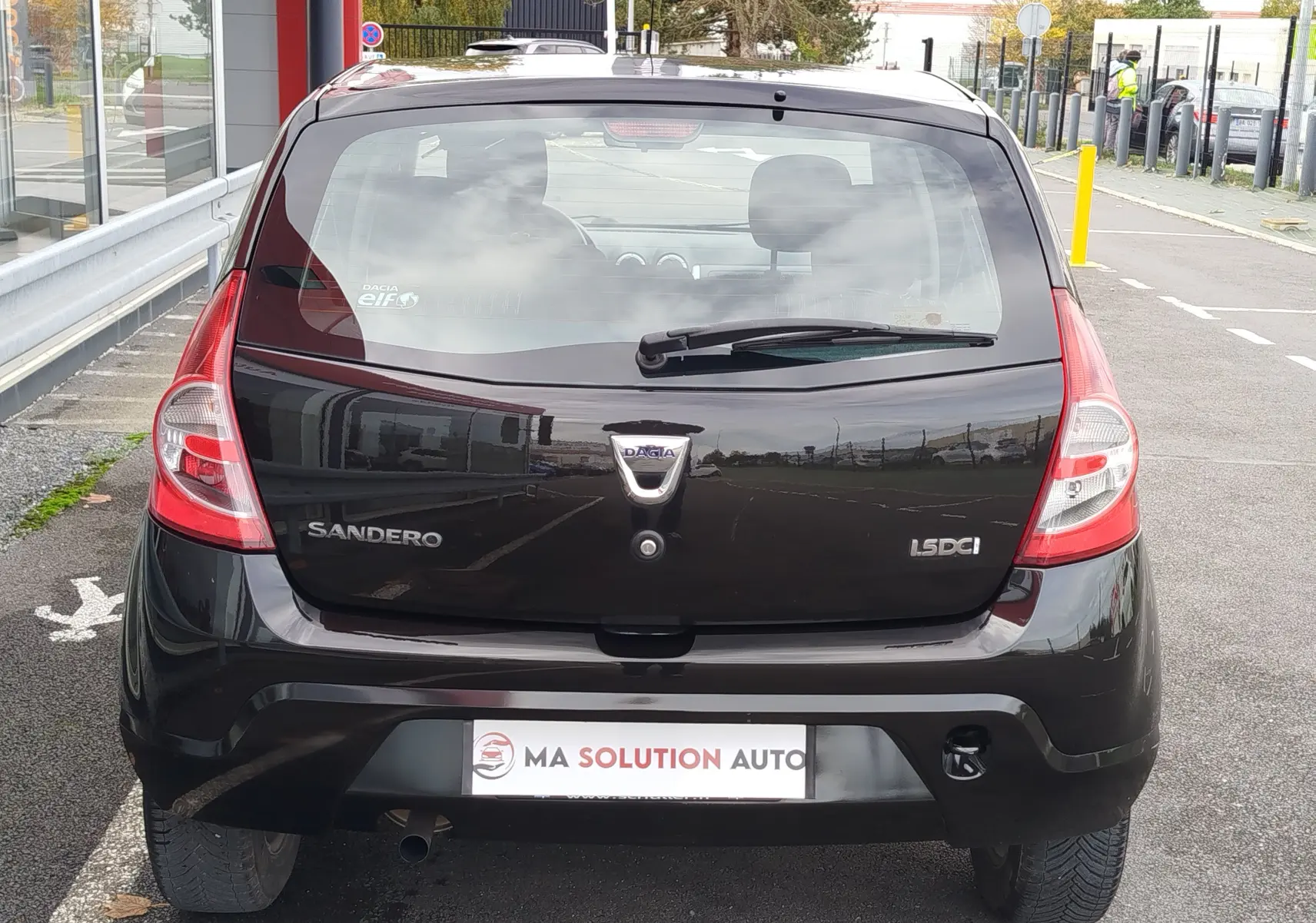 Vue arrière d'une Dacia Sandero noire de 2011, avec logo et badges 1.5 dCi visibles sur un parking urbain.