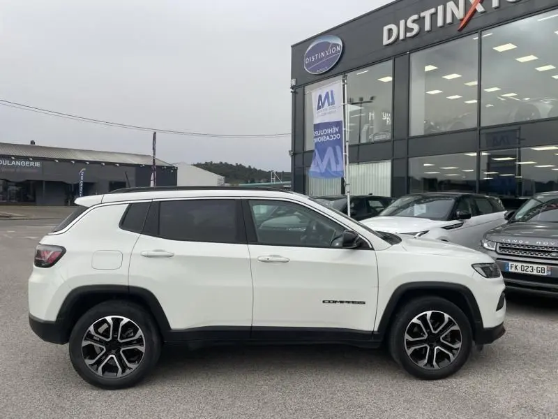 Vue de profil côté gauche d’un Jeep Compass blanc 2023 avec jantes alliage et vitres teintées, stationné devant un showroom.
