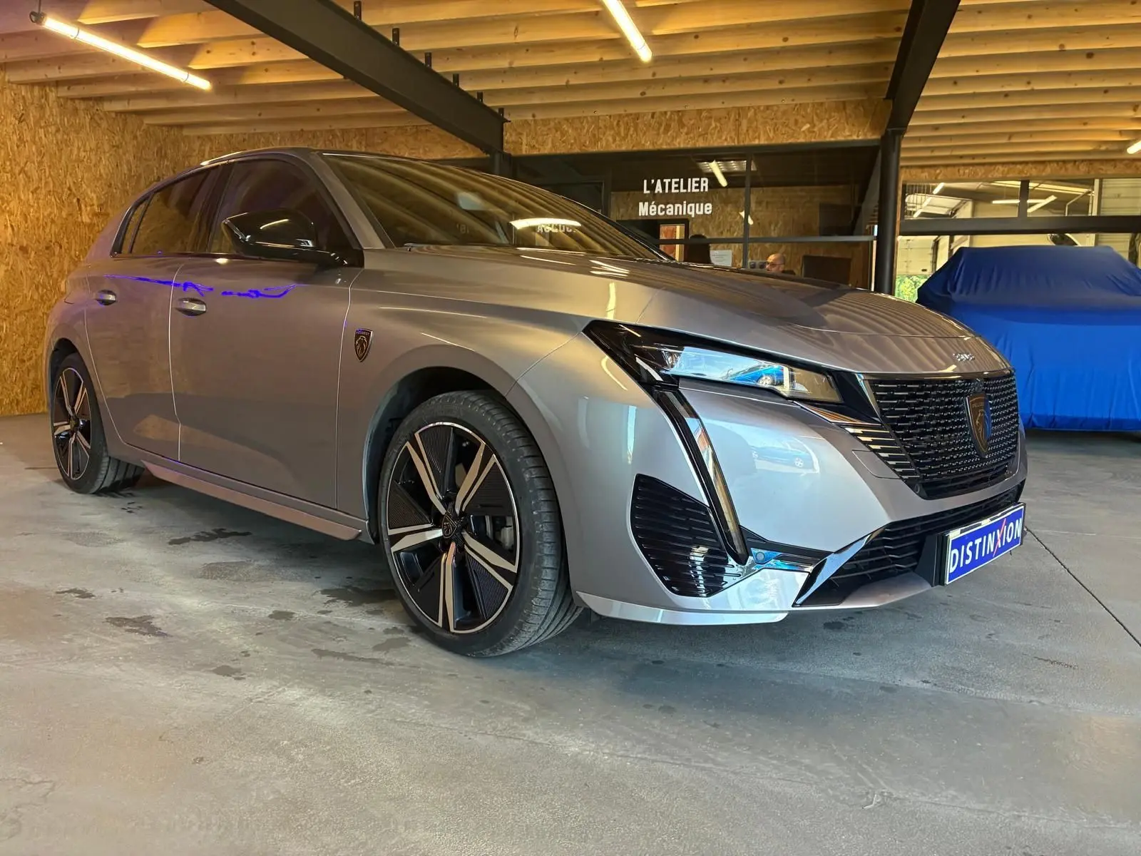 Peugeot 308 1.2 Hybrid gris vue 3/4 avant droit, avec jantes noires et calandre au design moderne dans un atelier.