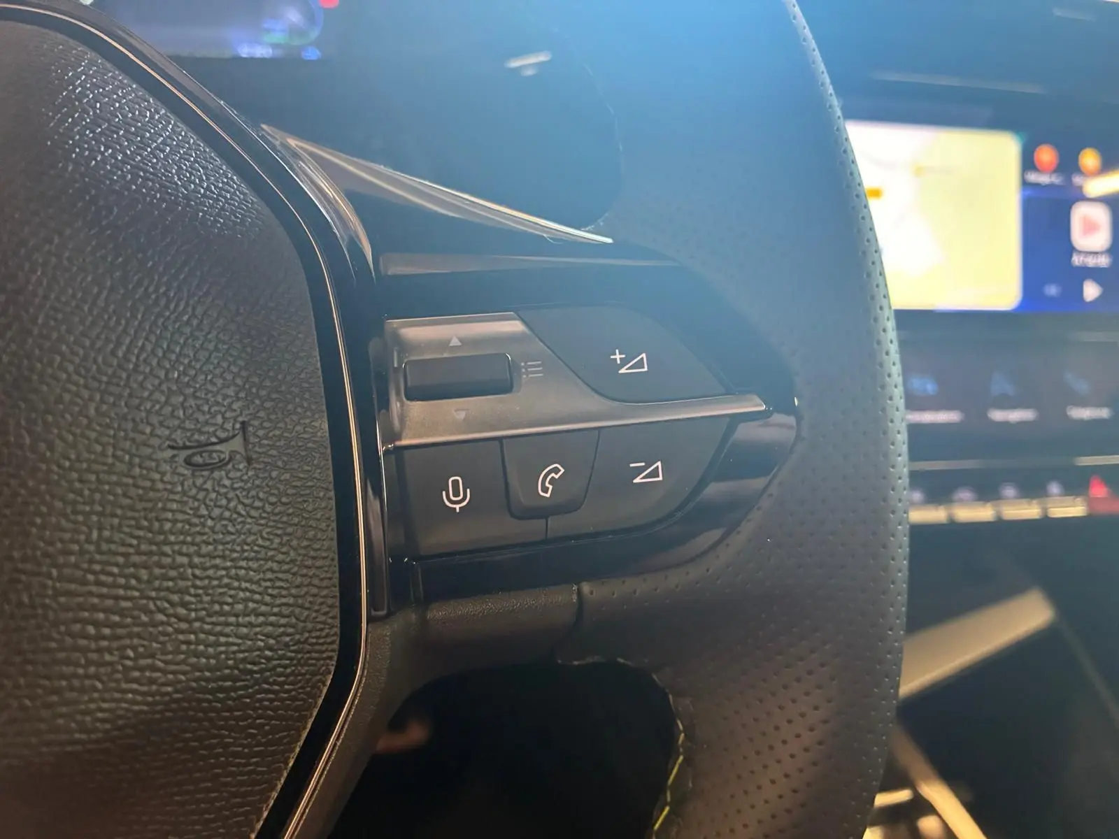 Gros plan sur les commandes gauche du volant perforé noir de la Peugeot 308 gris, avec écran tactile flou en arrière-plan.