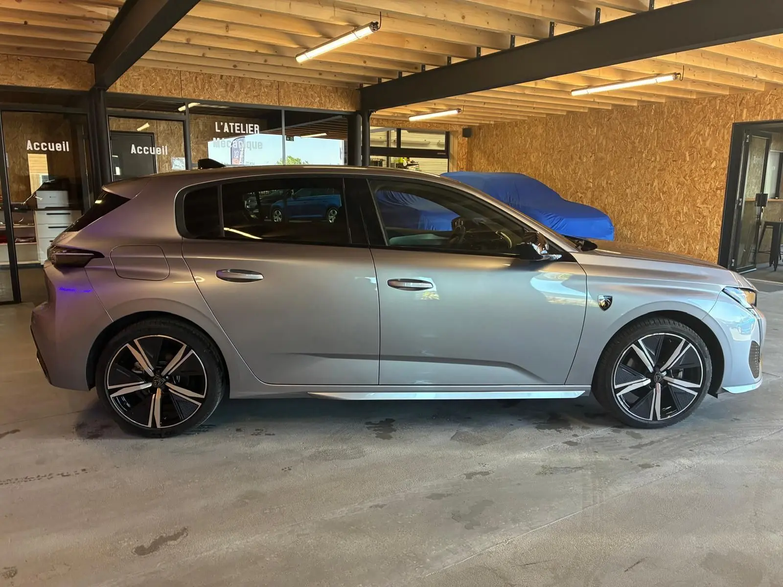 Profil droit d'une Peugeot 308 1.2 Hybrid 136 gris, avec jantes noires et détails sportifs, stationnée en intérieur.