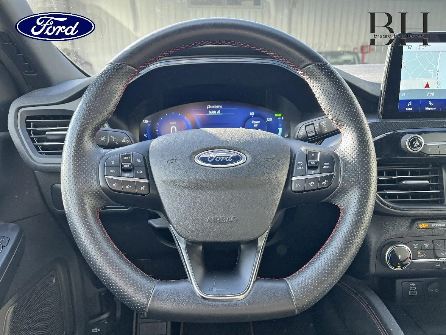 Vue frontale du volant cuir perforé noir avec surpiqûres rouges du Ford Kuga 2023, tableau de bord numérique et écran tactile GPS visible.