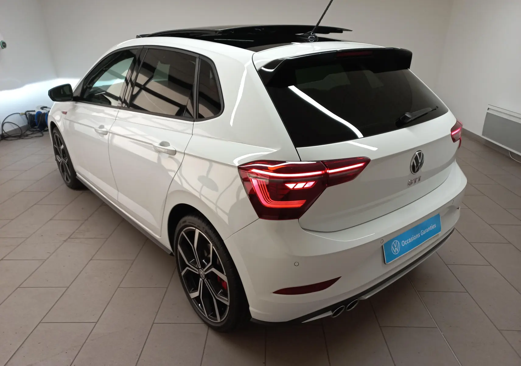 Vue 3/4 arrière droite d'une Volkswagen Polo GTI 2024 blanche avec toit noir ouvrant et feux arrière allumés.
