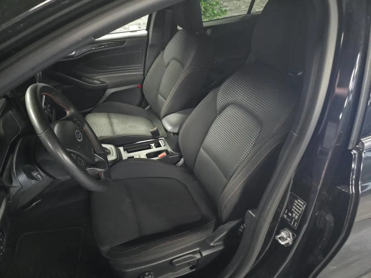 Vue intérieure côté conducteur de la Ford Focus noire 2024, sièges tissu ST-Line avec surpiqûres rouges et volant sport.