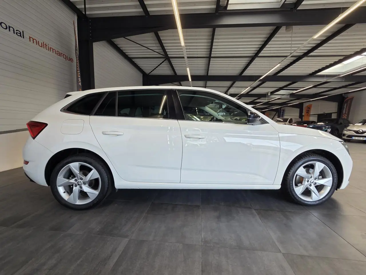 Vue latérale droite d'une Skoda Scala blanche 2020 avec jantes alliage argent et vitres teintées en intérieur showroom.