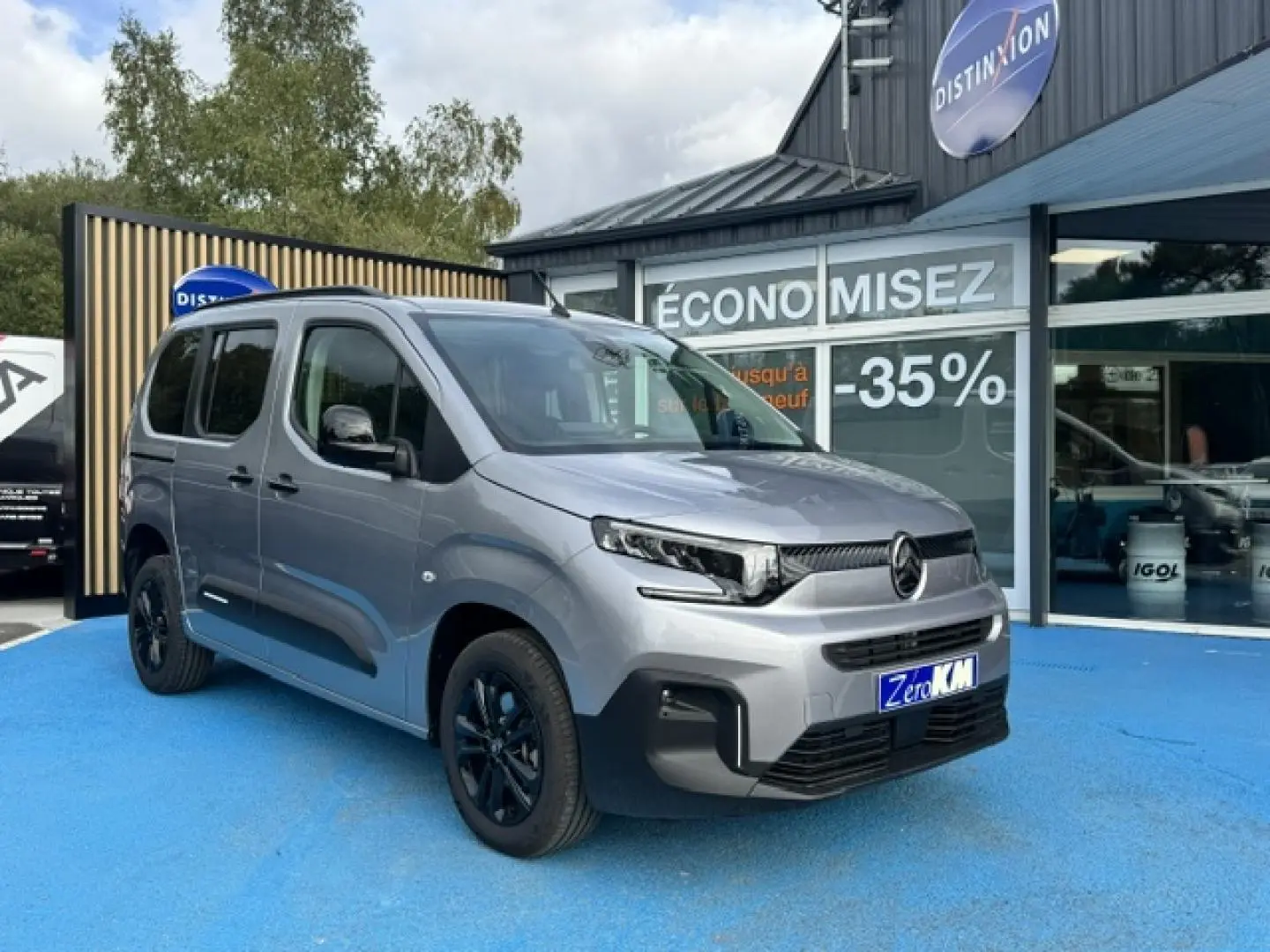 Vue 3/4 avant gauche d'un Citroën Berlingo gris acier avec jantes noires, stationné devant un magasin.