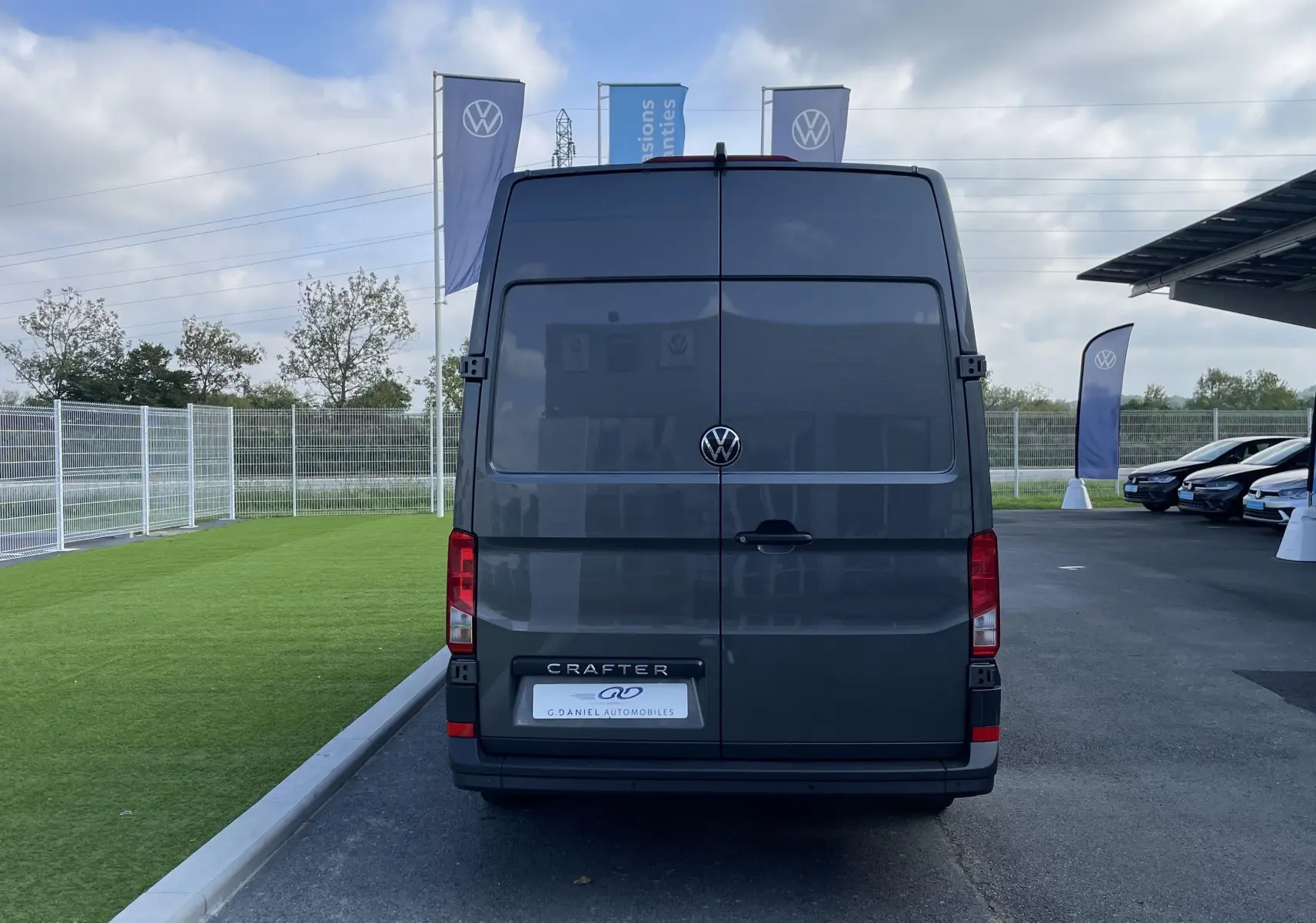 Vue arrière d'un Volkswagen Crafter Van gris Indium garé sur un parking extérieur avec drapeaux VW en arrière-plan.