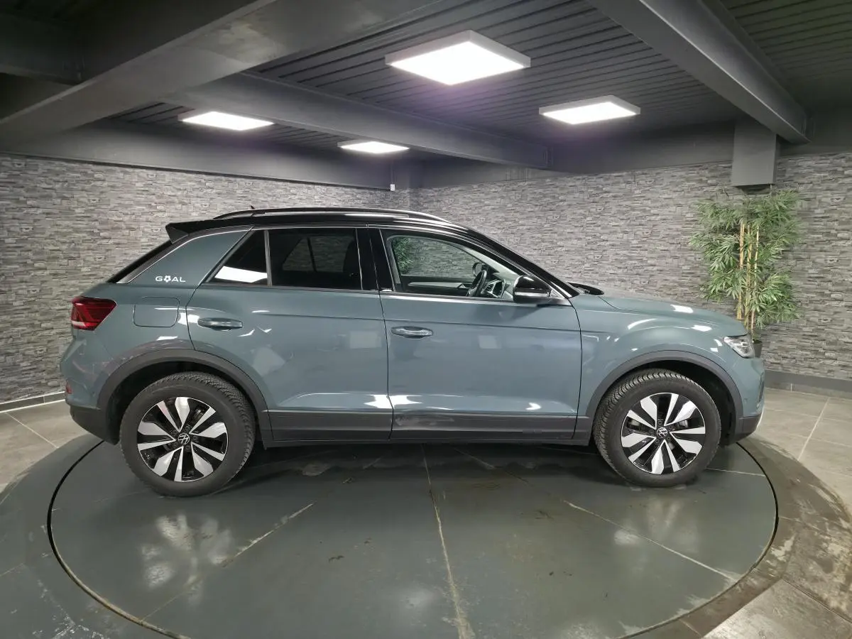Vue de profil côté gauche d'un Volkswagen T-Roc bleu métallisé avec toit noir et jantes distinctives en intérieur showroom.