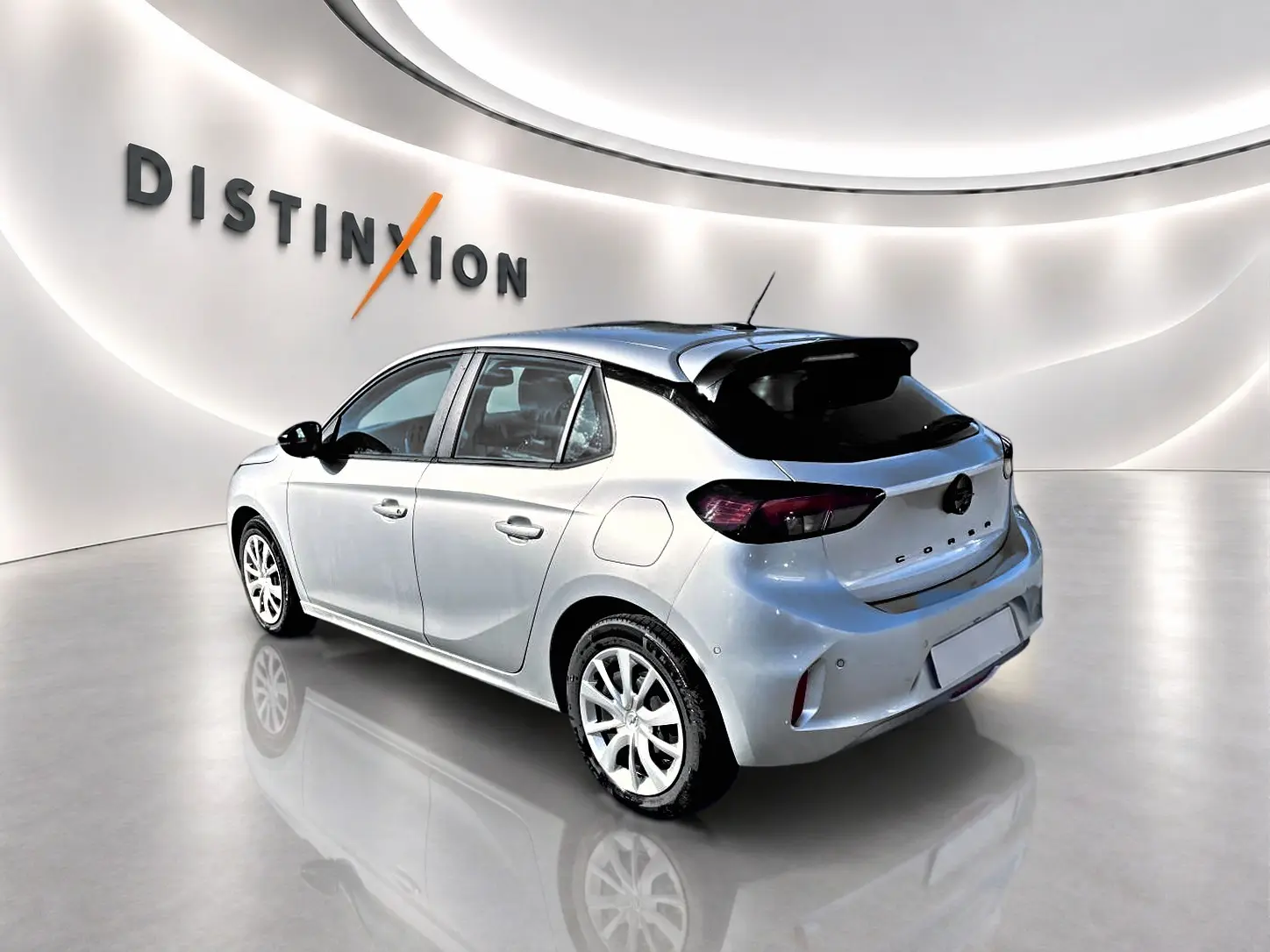 Opel Corsa gris 3/4 arrière droit en intérieur, avec becquet noir et jantes blanches distinctives.