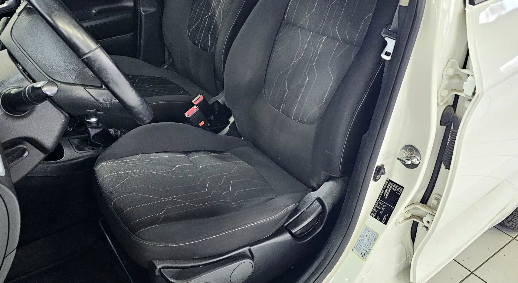 Vue intérieure côté conducteur d'une Kia Picanto II blanche de 2016, sièges noirs avec surpiqûres et volant visible.