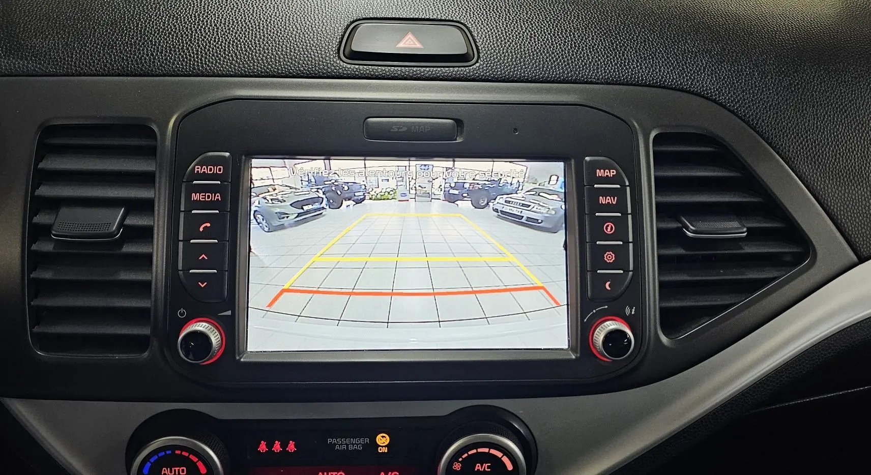 Écran central de la KIA PICANTO blanche 2016 montrant la caméra de recul avec les lignes de guidage colorées.