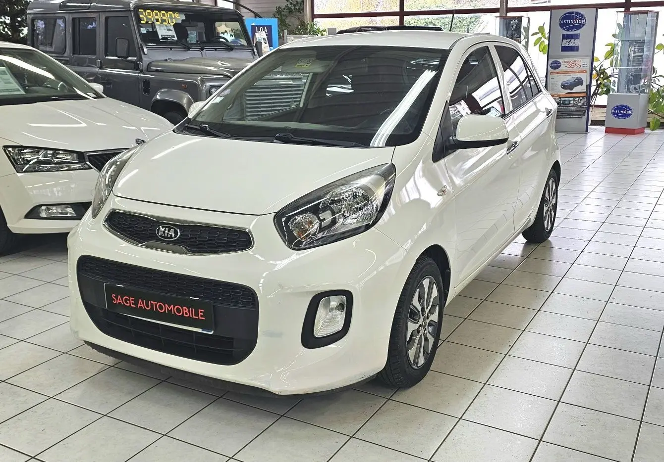 KIA Picanto blanc vue 3/4 avant droit en showroom avec plaque SAGE AUTOMOBILE visible