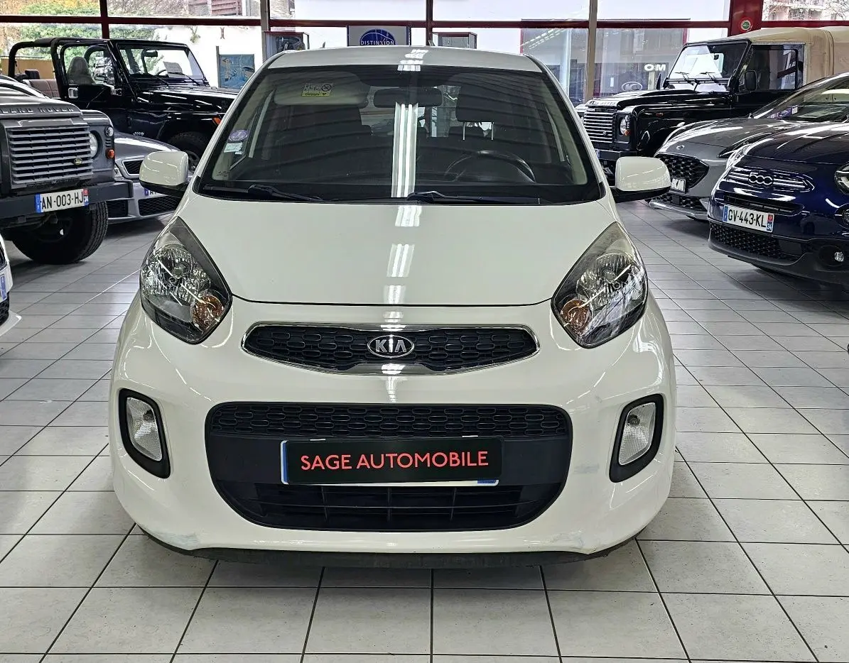 Vue avant d'une KIA PICANTO II blanche de 2016 en showroom, avec calandre noire et phares arrondis visibles.
