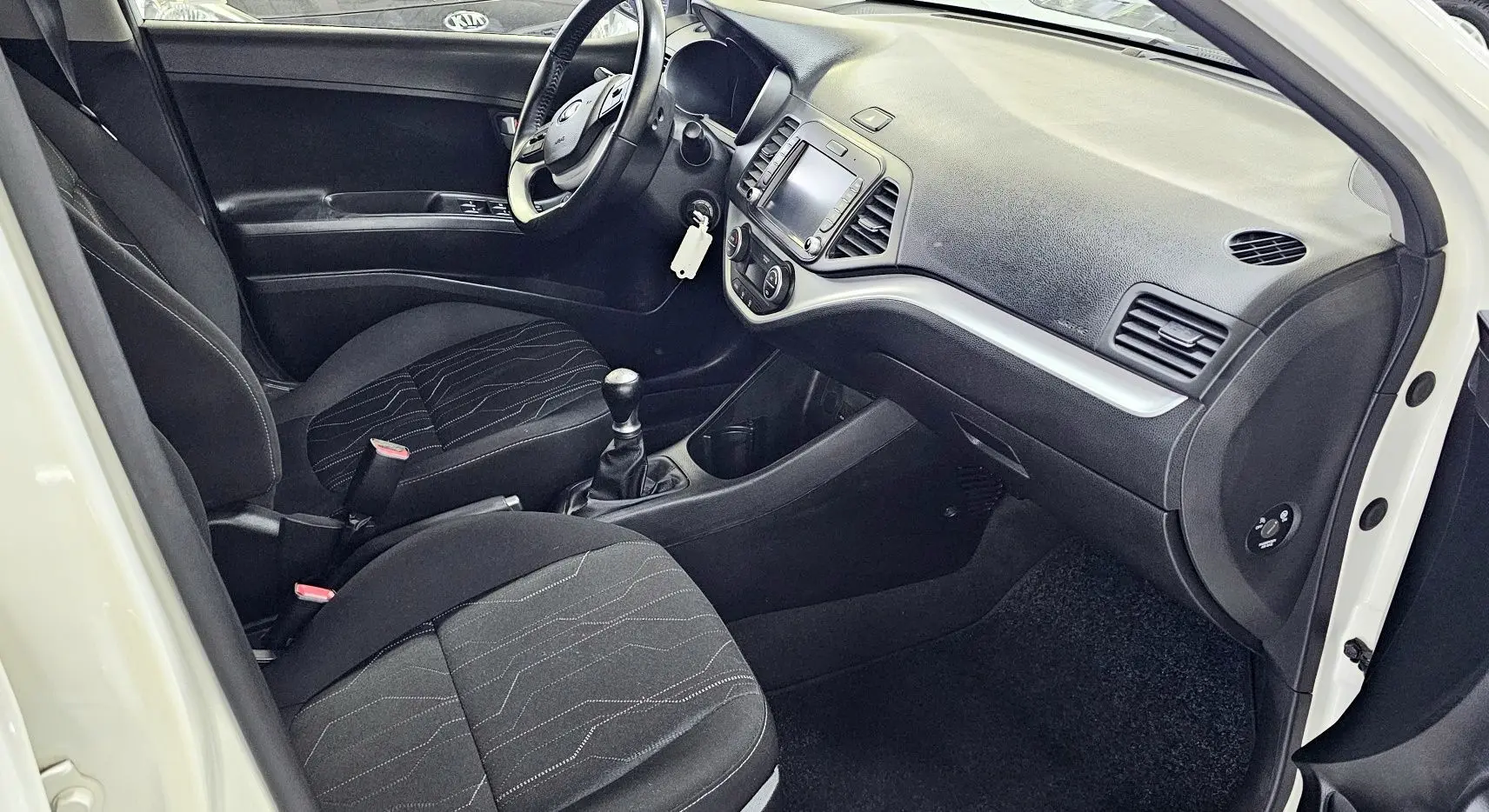 Intérieur noir de la Kia Picanto II blanche 2016, vue côté conducteur avec tableau de bord et boîte manuelle visibles.