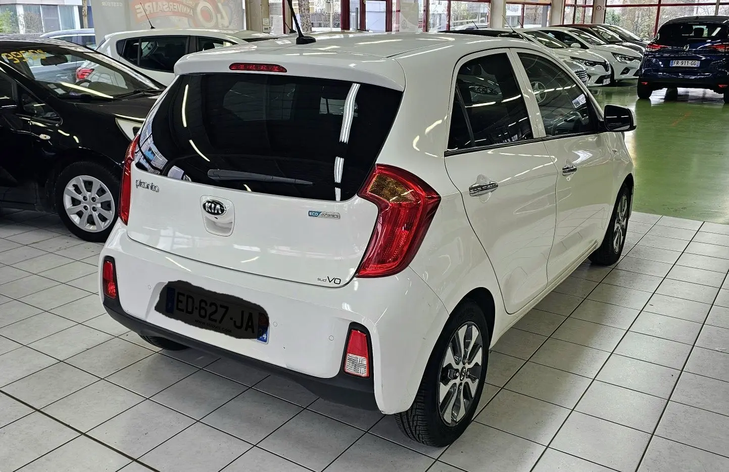 Vue 3/4 arrière droite d'une KIA Picanto blanche 2016 en showroom, avec feux arrière rouges et vitres teintées.