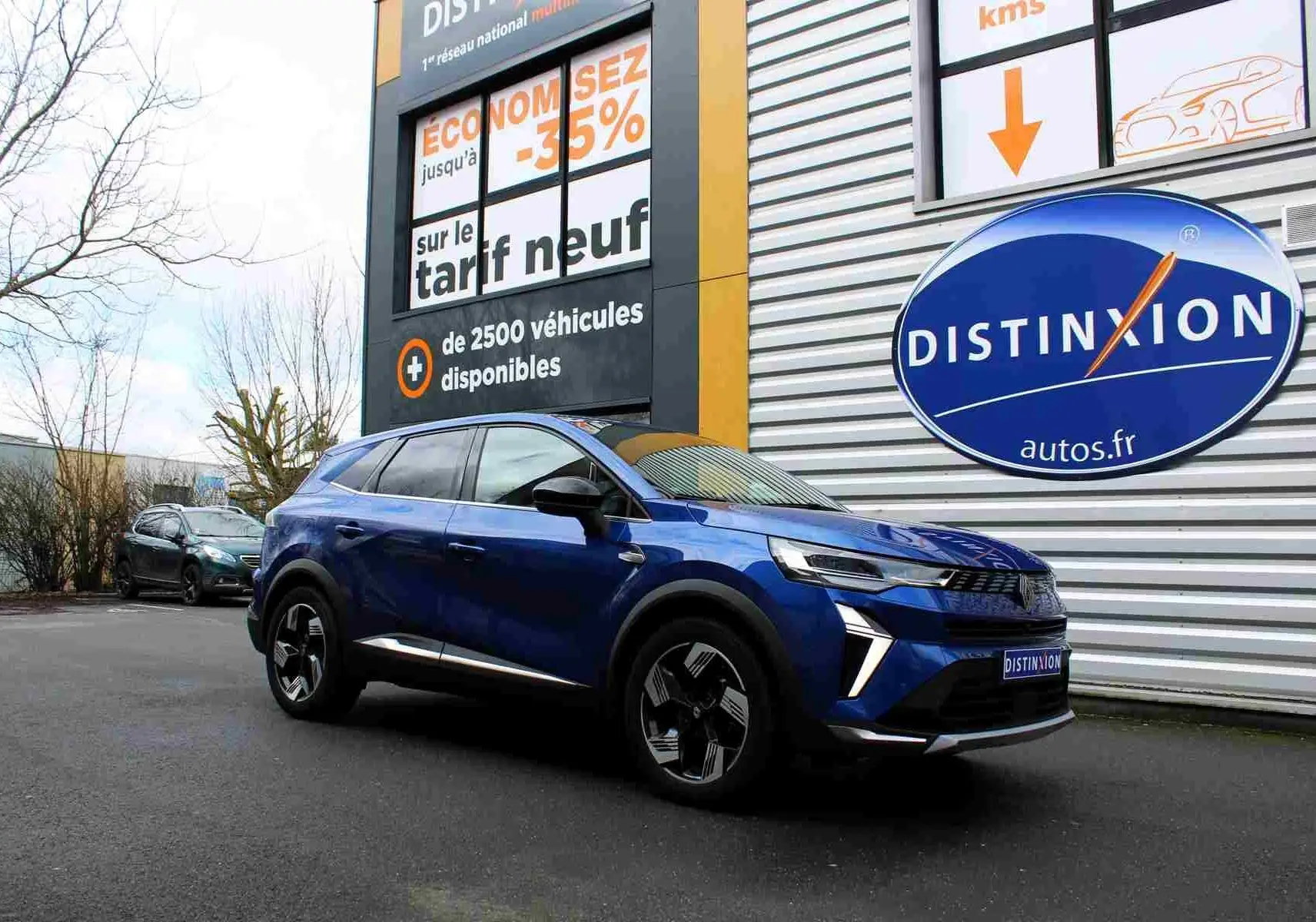 Renault Symbioz bleu Iron Metal en 3/4 avant droit, avec calandre noire et jantes au design futuriste, devant un bâtiment commercial.