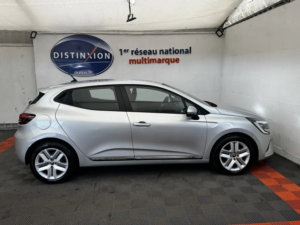 Profil côté gauche d'une Renault Clio Business E-TECH 140 gris clair, avec jantes alliage et toit spoiler visible en intérieur showroom.