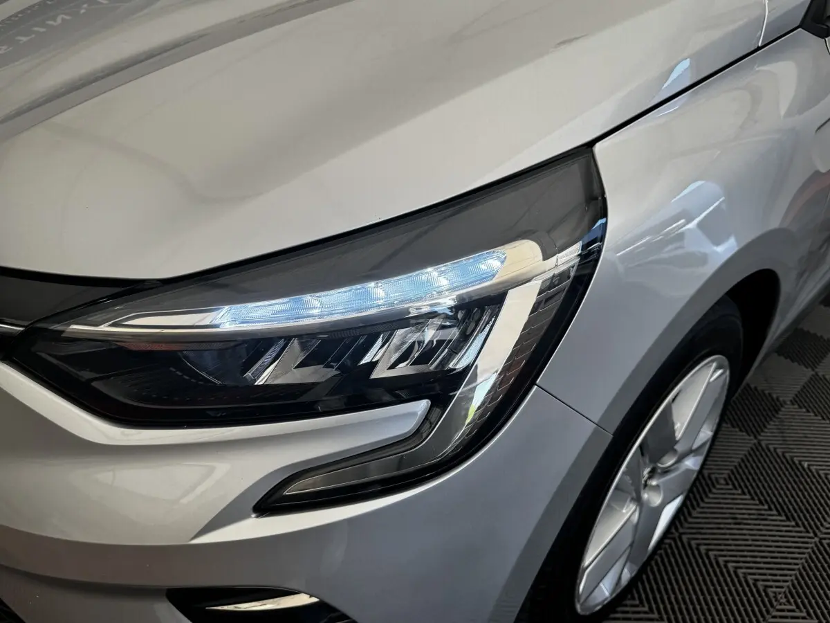 Gros plan sur le phare avant droit allumé de la Renault Clio Business E-TECH 140 gris clair, avec jante visible.