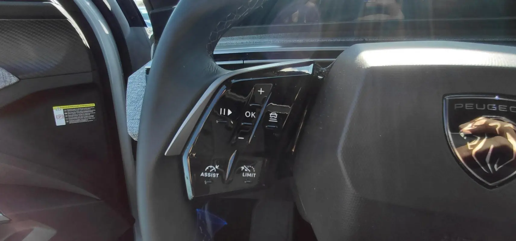 Gros plan sur les commandes gauche du volant noir du Peugeot 5008 Hybrid blanc 2025, avec logo lion visible.