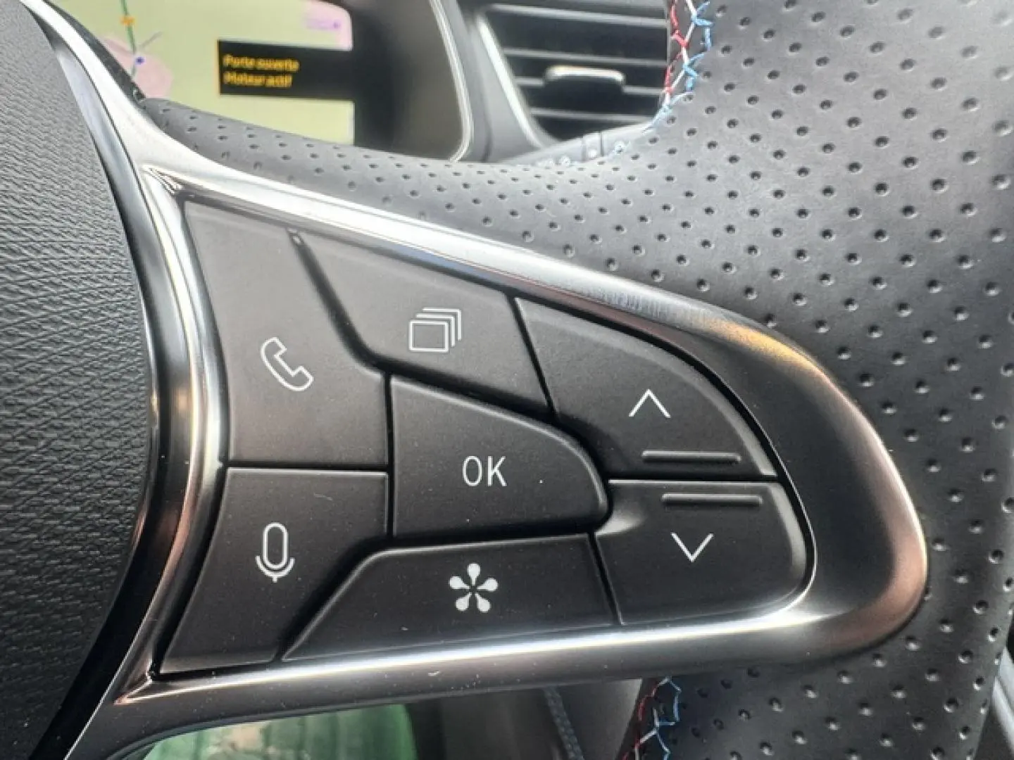 Gros plan sur les commandes noires du volant perforé de la Renault Symbioz gris Cassiopée, avec surpiqûres bleues.