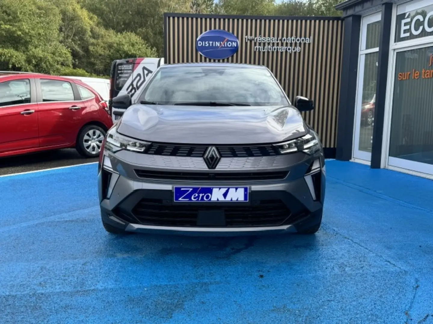 Vue avant d'une Renault Symbioz gris Cassiopée métallisé stationnée sur un sol bleu devant un bâtiment commercial.