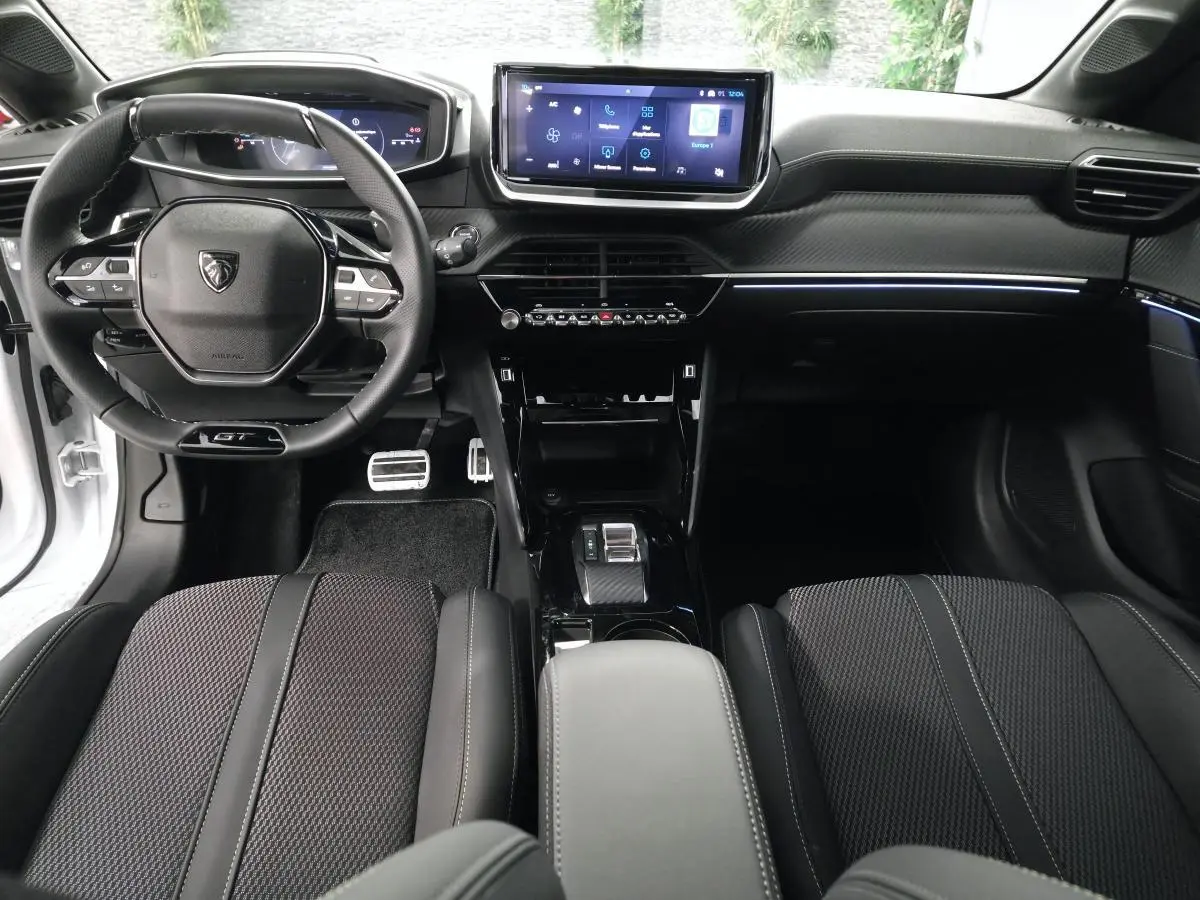 Vue intérieure avant de la Peugeot 208 blanche 2025, volant compact GT, écran tactile central et sellerie noire.