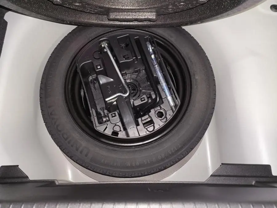 Vue plongeante sur la roue de secours galette noire et ses outils dans le coffre blanc d'une Peugeot 208 1.2i Hybrid 2025.