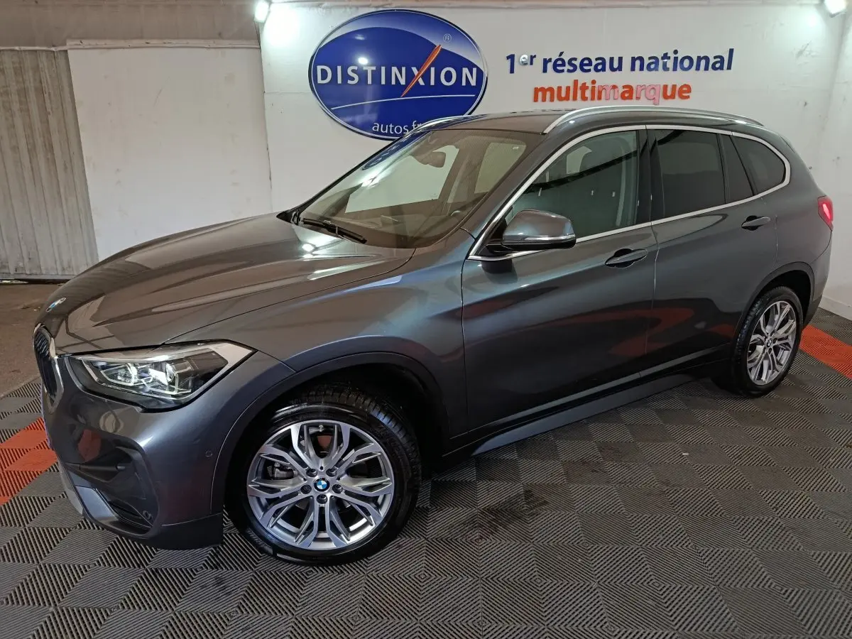 BMW X1 sDrive18i gris foncé vue 3/4 avant droit, feux allumés, roues alliage et rails de toit noirs visibles