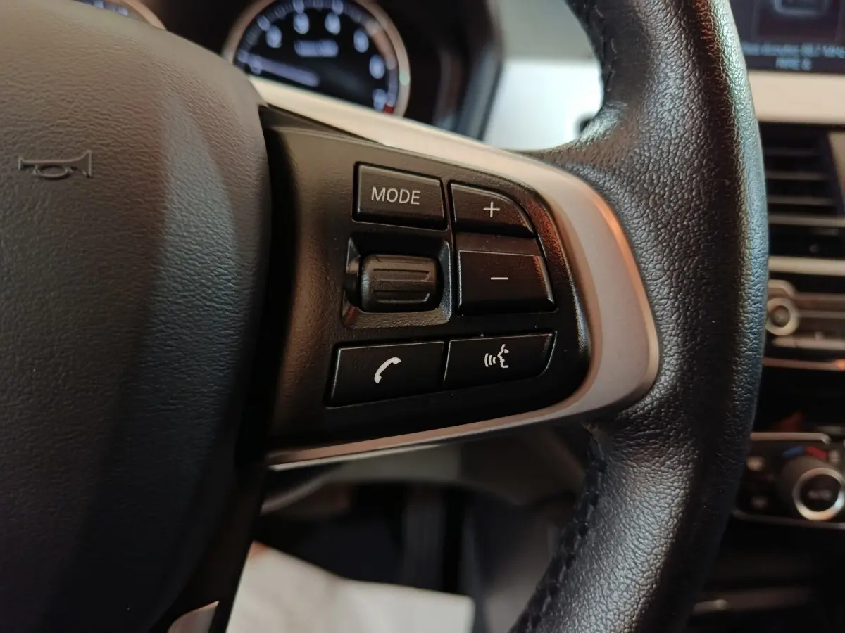 Gros plan sur les commandes gauche du volant cuir noir du BMW X1 sDrive18i Lounge 2021, avec tableau de bord flou en arrière-plan.