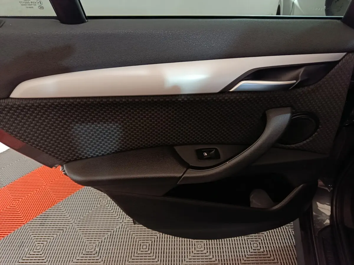 Vue intérieure du panneau de porte côté gauche d'une BMW X1 sDrive18i Lounge gris foncé, avec poignée et bouton de verrouillage.