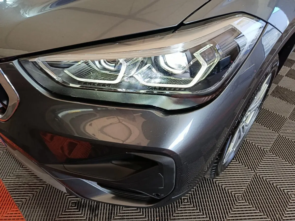 Gros plan sur le phare avant droit d’un BMW X1 sDrive18i Lounge gris foncé avec jante et partie du pare-chocs visibles.