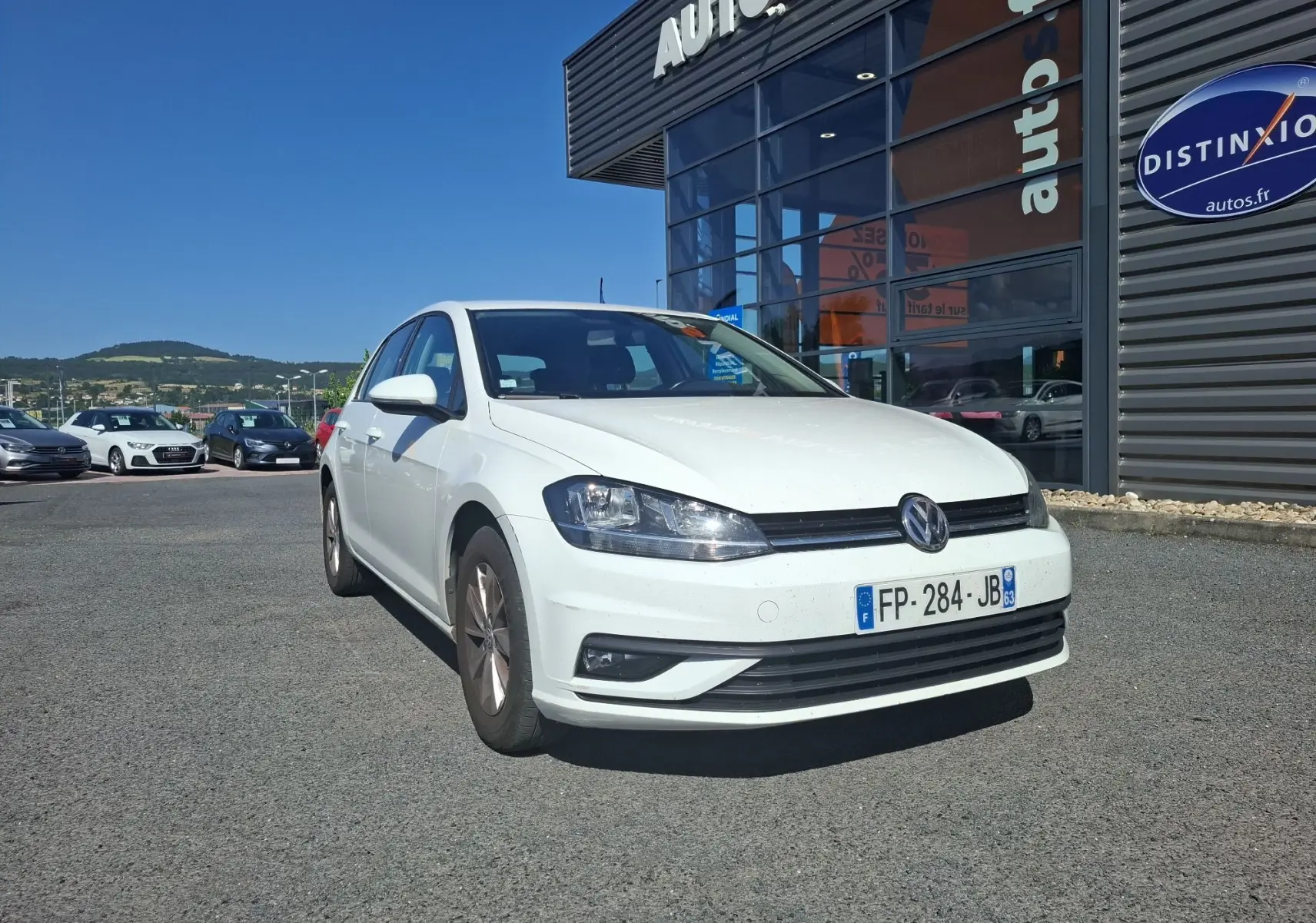 Volkswagen Golf blanc vue 3/4 avant droit, garée devant un bâtiment commercial sous un ciel bleu.