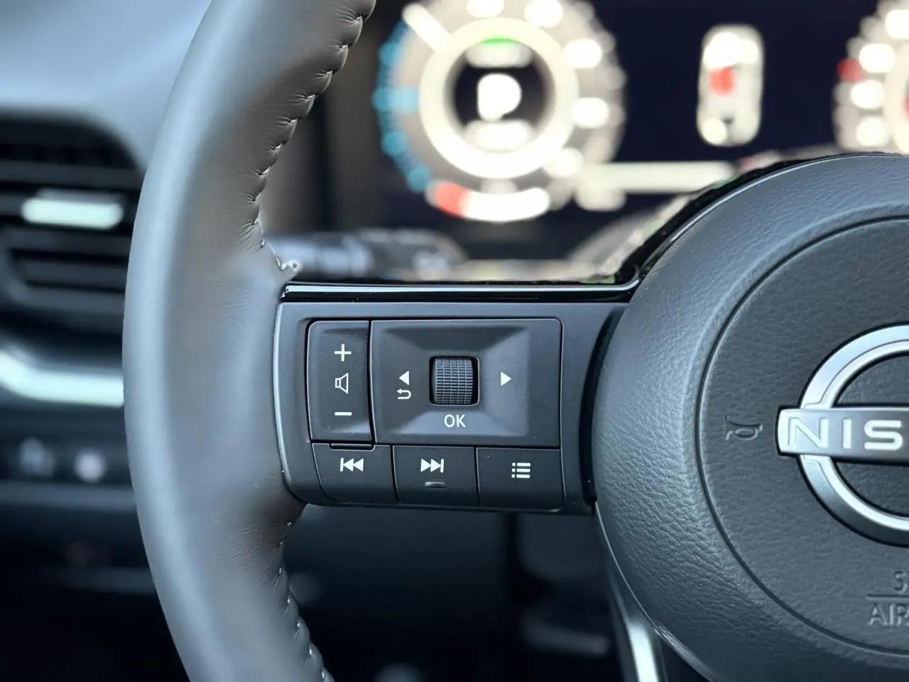 Gros plan sur les commandes gauche du volant cuir du Nissan Qashqai gris Ceramic, avec tableau de bord numérique flou en arrière-plan.