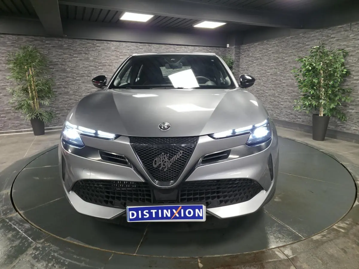 Vue avant d'une Alfa Romeo Junior 1.2 Ibrida gris métallisé avec calandre noire et phares LED allumés.