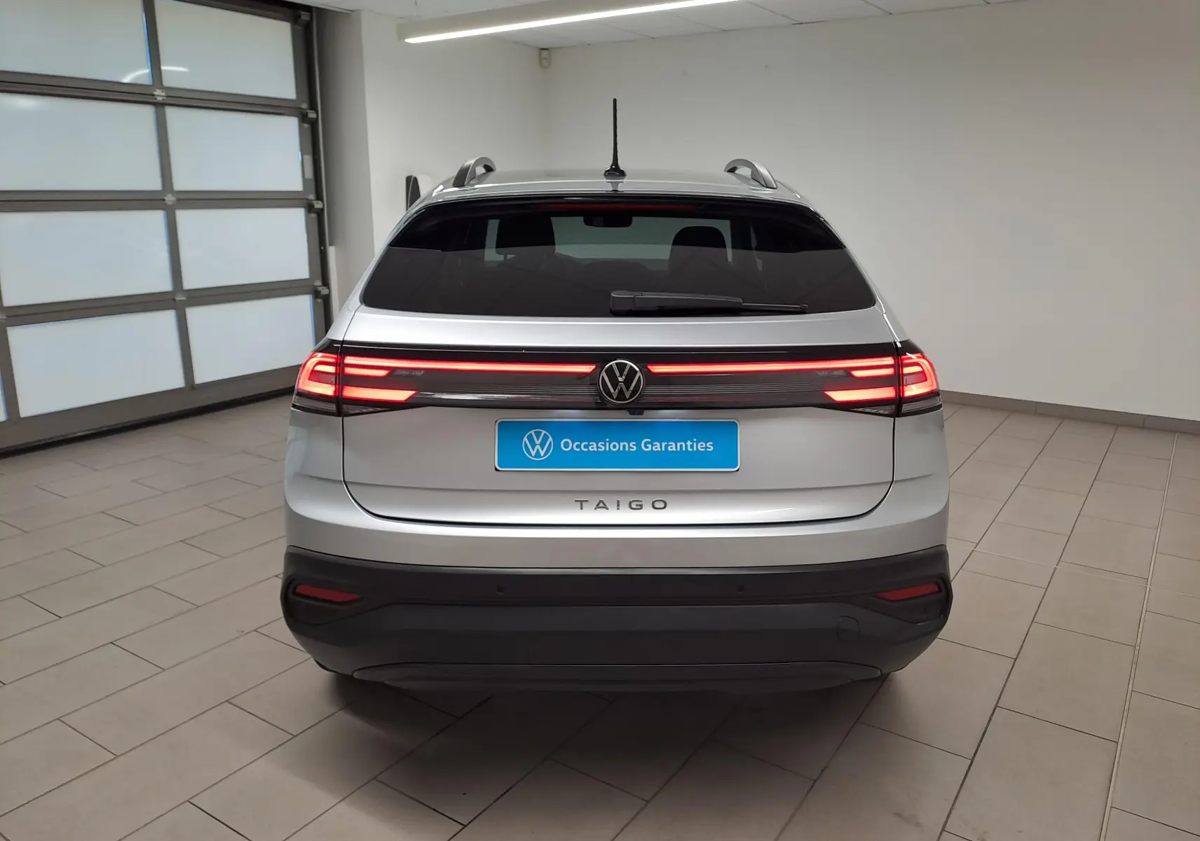 Vue arrière du Volkswagen Taigo 2025 en peinture métallisée Reflet d'argent avec feux LED allumés dans un garage.