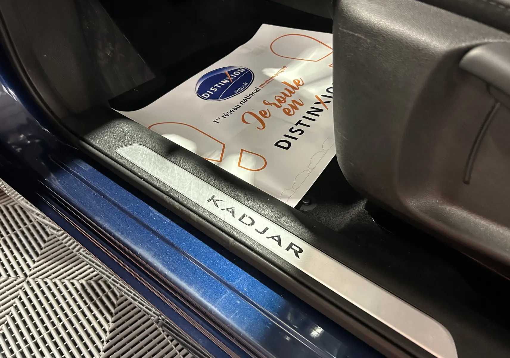Gros plan sur la plaque de seuil de porte côté gauche du Renault Kadjar bleu, avec tapis de sol et siège noir visible.