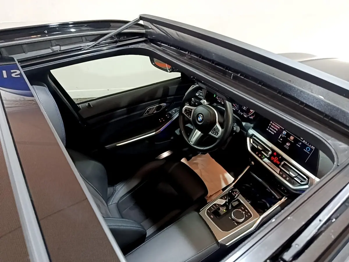 Vue intérieure par le toit ouvrant d'une BMW Série 3 noire, mettant en valeur le tableau de bord digital et le volant cuir.