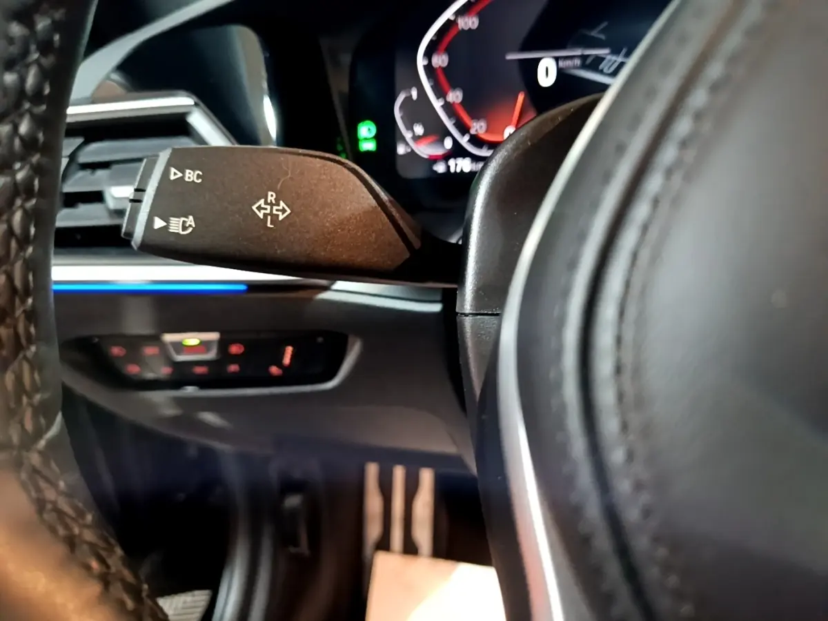 Gros plan sur la manette de clignotant à gauche du volant cuir d'une BMW Série 3 318i noire, tableau de bord numérique en arrière-plan.