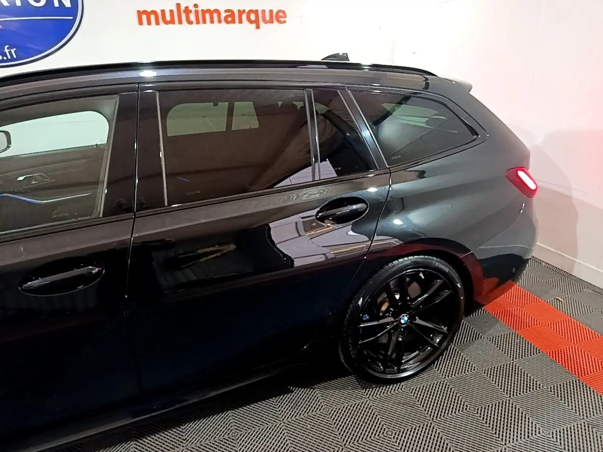 BMW Série 3 318i noire vue de profil côté gauche, avec jantes noires et feu arrière allumé.