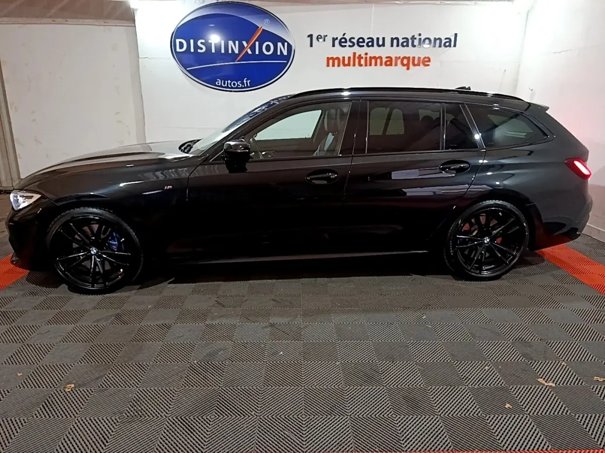 BMW Série 3 318i M Sport noir vue de profil côté gauche, avec jantes noires et feux arrière allumés dans un garage.