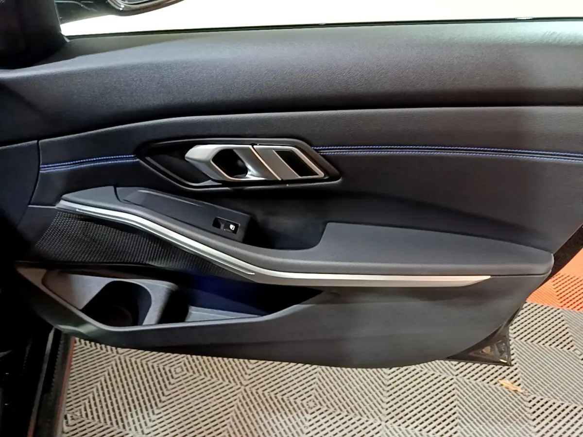 Gros plan sur la porte côté gauche noire de la BMW Série 3 318i M Sport 2022 avec surpiqûres bleues et poignée alu.