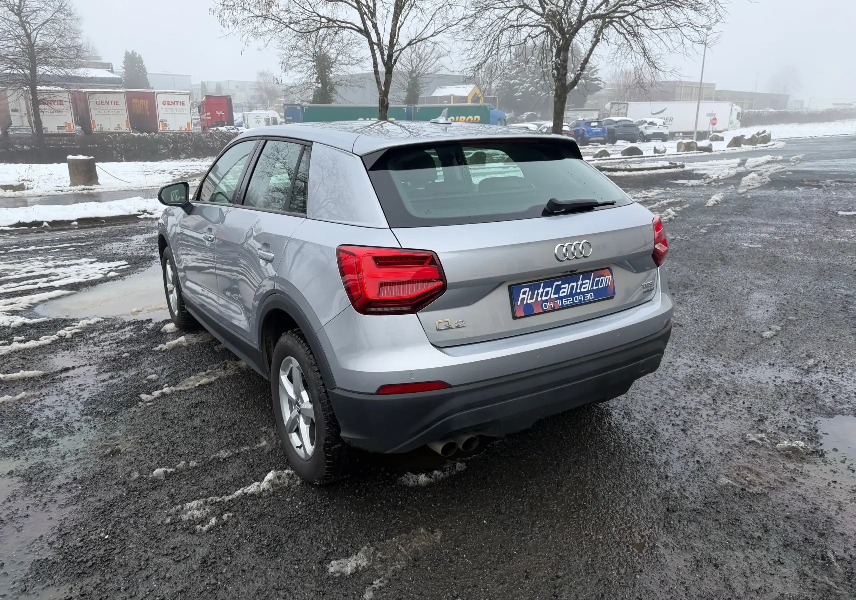 Audi Q2 gris aluminium vue 3/4 arrière droit sur sol humide avec neige et feux arrière rouges visibles.