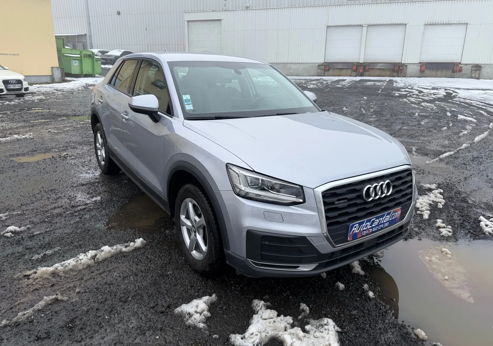 Audi Q2 gris aluminium vue 3/4 avant droit sur sol humide avec traces de neige et boue.