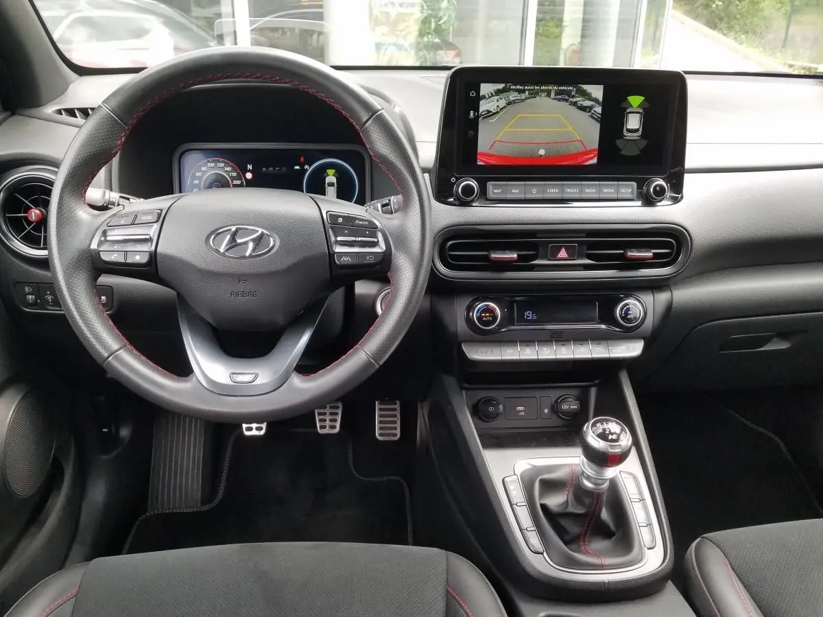 Vue intérieure avant du Hyundai Kona rouge 2023, volant cuir, écran tactile avec caméra de recul et boîte manuelle.