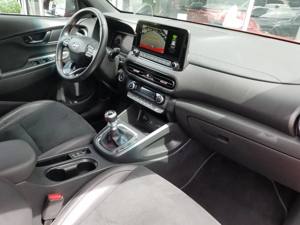 Vue intérieure côté conducteur du Hyundai Kona rouge 2023, avec tableau de bord numérique et levier de vitesse manuel.