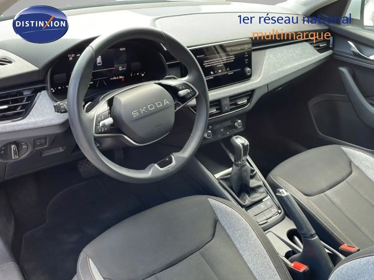 Vue intérieure centrée sur le volant et la console centrale d'une Skoda Scala blanche Glacier avec sellerie tissu gris noir.