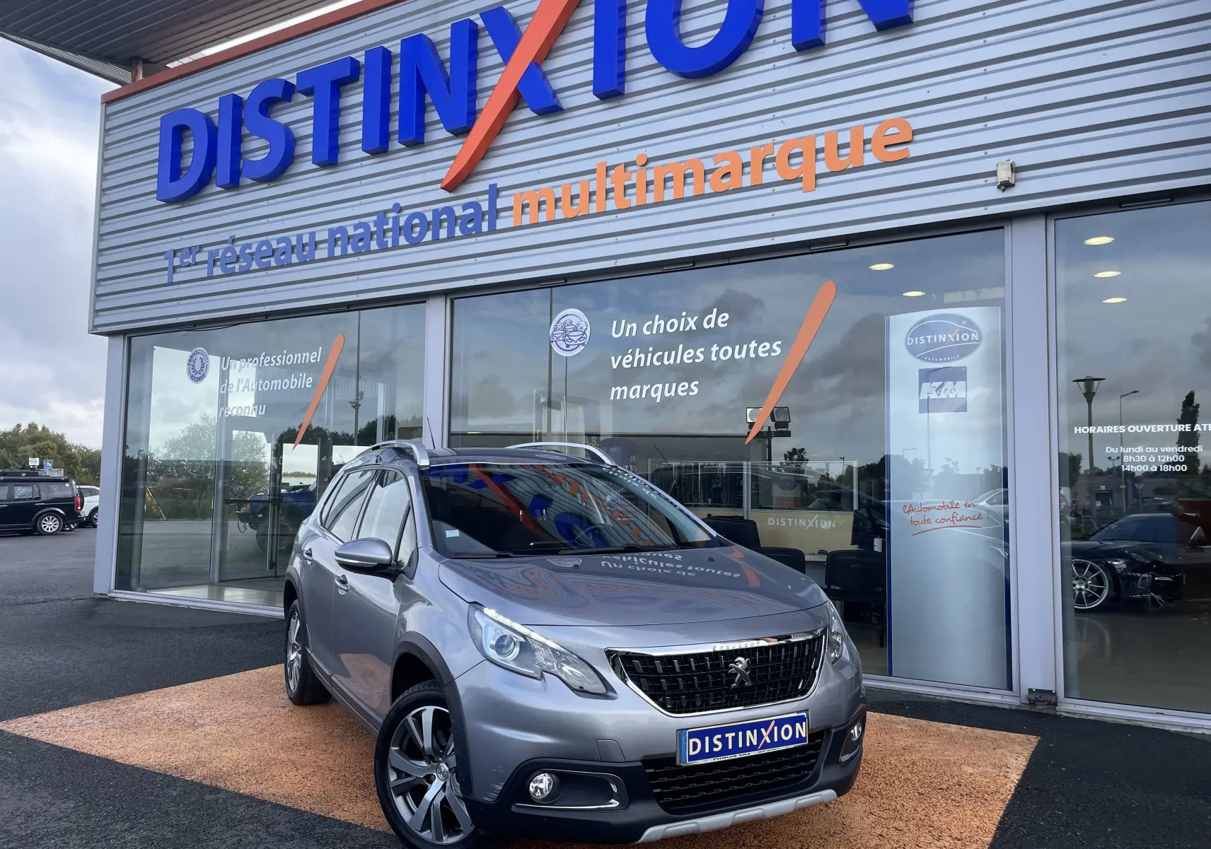Peugeot 2008 gris en 3/4 avant droit devant une concession Distinxion, avec jantes alliage et calandre noire.